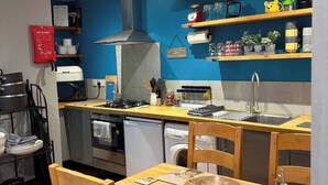 A compact kitchen with a stove, oven, and sink, wooden chairs, a dining table, and blue walls. - The Barn at The Flute - Mill Meece (Stafford)