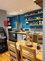 Una cocina compacta con estufa, horno y lavamanos, sillas de madera, una mesa de comedor y paredes azules.