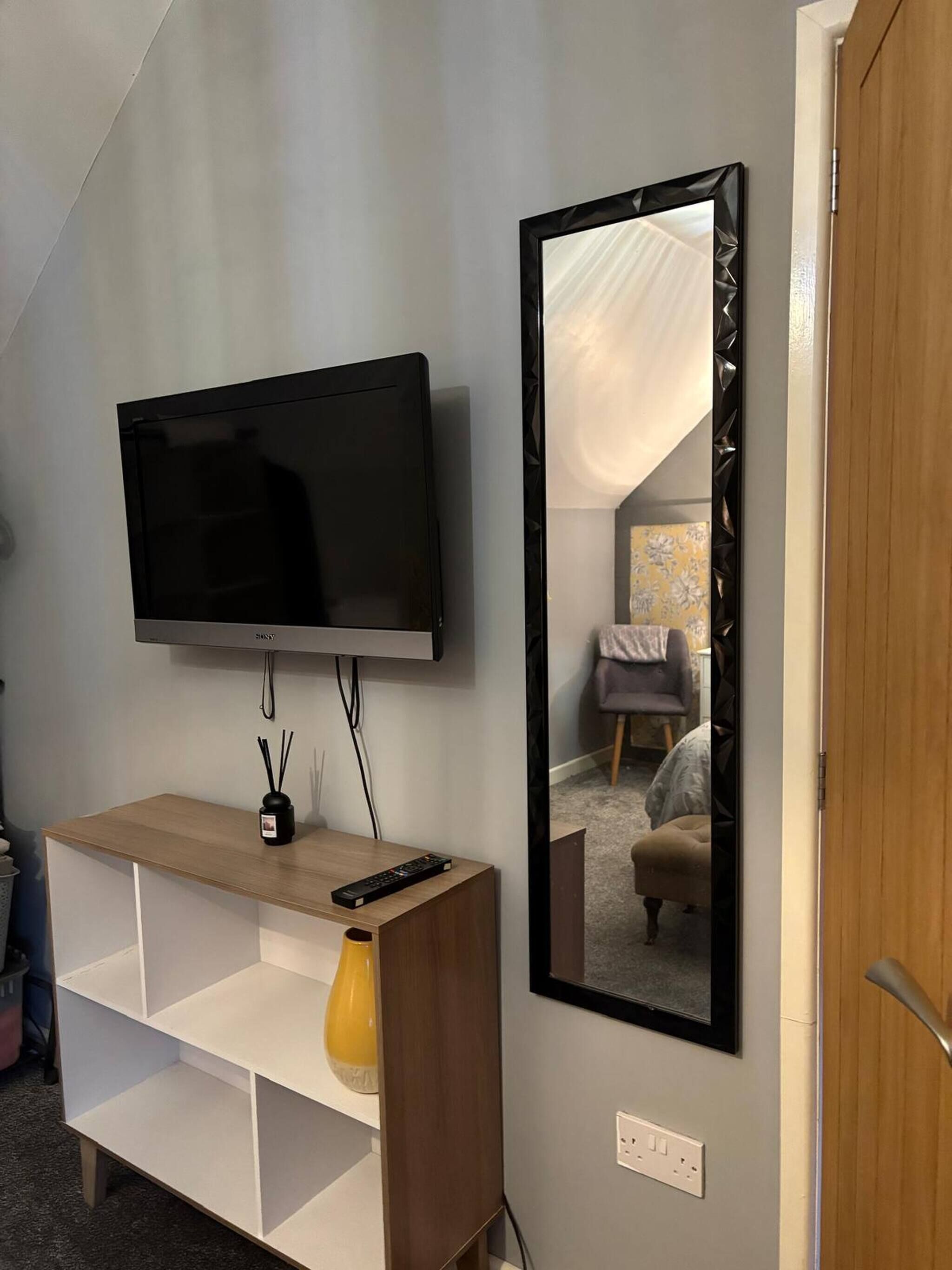 A modern hotel room with a flat-screen TV mounted on the wall, a wooden console table with a remote control, a mirror, and a small yellow vase.