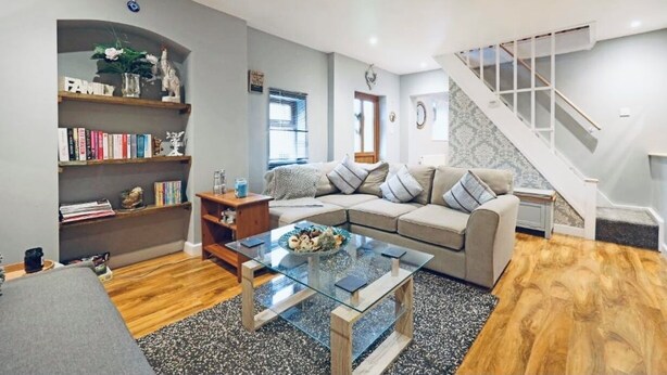 A living room with a glass coffee table, a beige sofa, a bookshelf, and a staircase. - The Barn at The Flute - Mill Meece (Stafford)