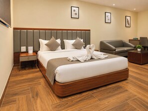 A hotel room with a large bed, a wooden nightstand, a grey sofa, a small table, and a potted plant. - S by Z Hotels, Baani Square (Gurugram)