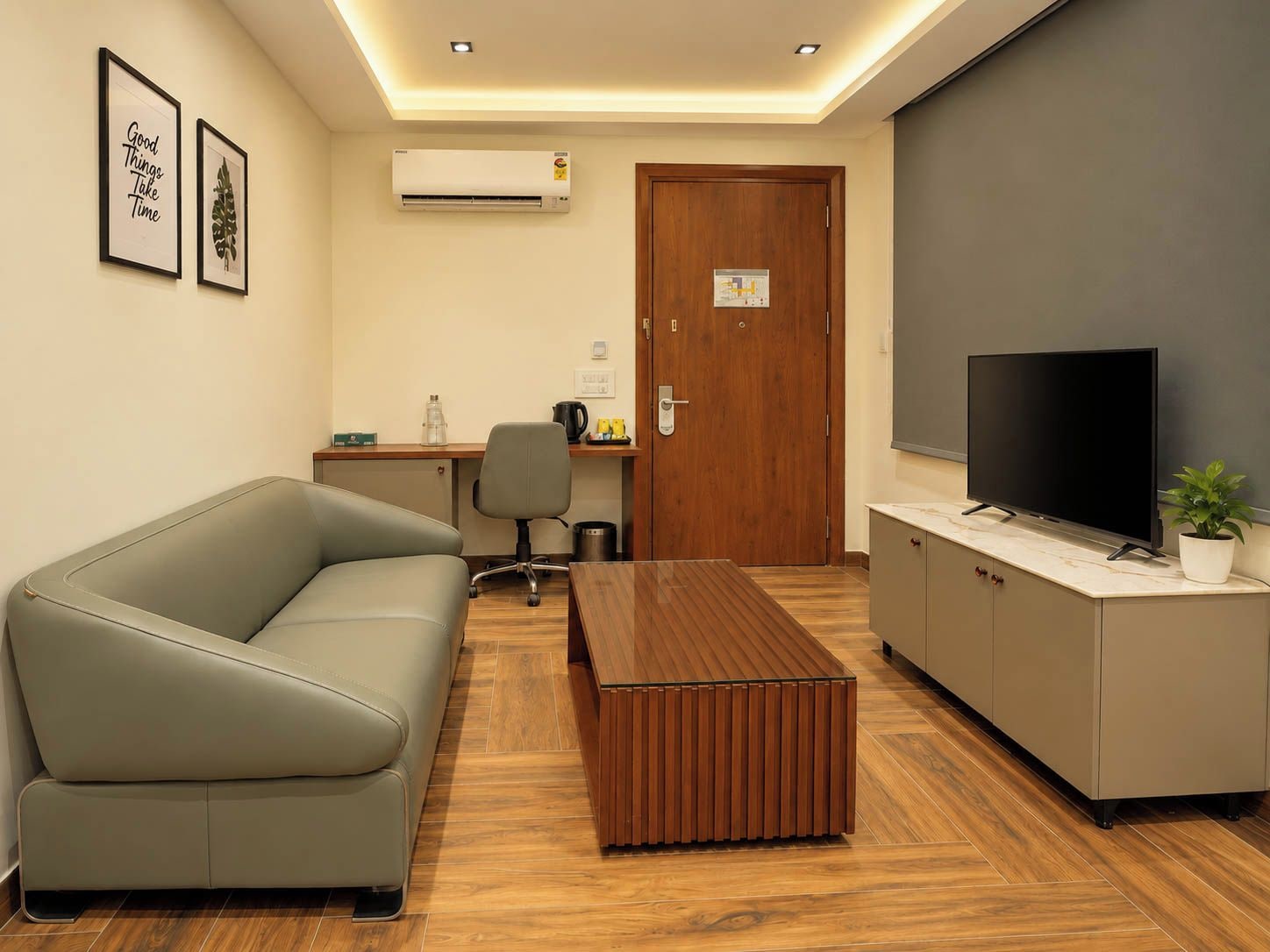 A modern office space with a sofa, a wooden coffee table, a desk with a chair, a television, and an air conditioning unit.