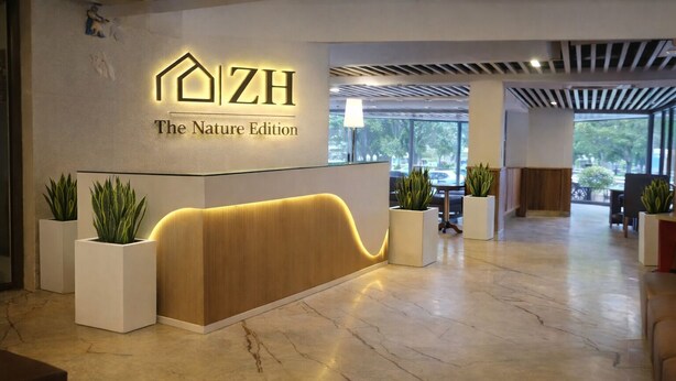 A reception desk with a modern design, labeled "ZH The Nature Edition." There are potted plants on either side of the desk, and the area is well-lit with natural light coming through large windows. - S by Z Hotels, Baani Square (Gurugram)