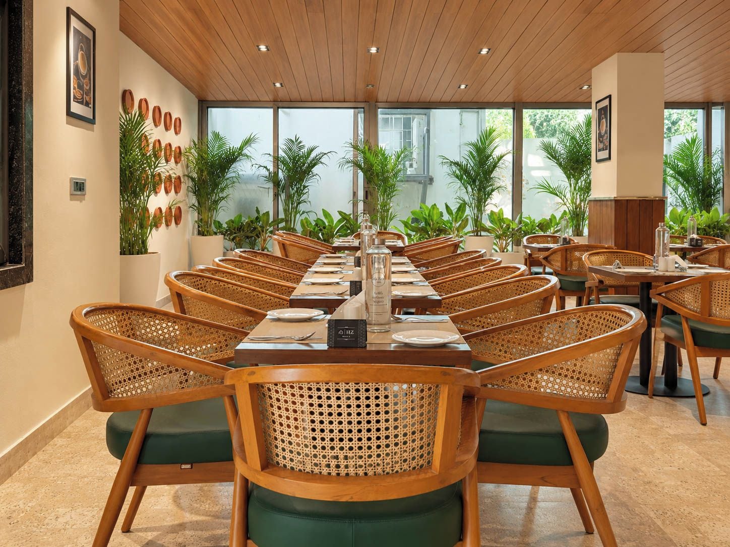 A long dining table with wicker chairs, surrounded by potted plants, in a well-lit room with wooden ceiling panels.