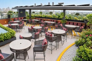 An outdoor patio with round tables and wicker chairs, surrounded by greenery and overlooking a cityscape. - S by Z Hotels, Baani Square (Gurugram)