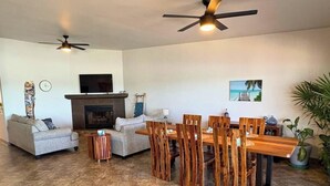 Uma sala de estar com lareira, dois sofás, uma mesa de jantar com cadeiras, ventiladores de teto e uma TV fixada na parede.