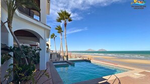 Uma área de piscina com vista para a praia e oceano, rodeada por palmeiras e um edifício com varandas.