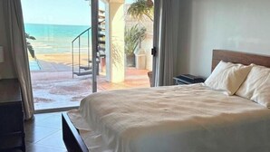 Quarto com cama, mesa de cabeceira, uma cadeira e vista para a praia por uma porta de correr.