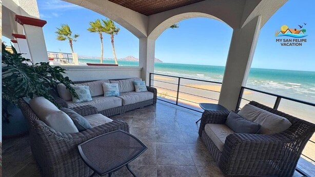 Um alpendre coberto com móveis de vime, uma mesa pequena e vista para a praia.