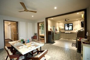 A spacious dining area with a large table and chairs, a kitchen with modern appliances, and a ceiling fan. - The Nivaas At Berhin's Farmstay (Ajmer)