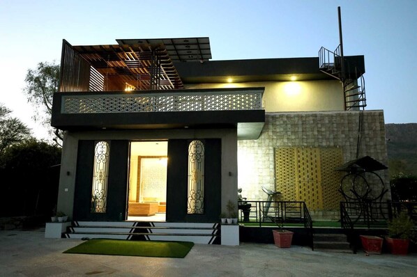 A modern two-story house with a balcony, a staircase, and a small garden area. - The Nivaas At Berhin's Farmstay (Ajmer)