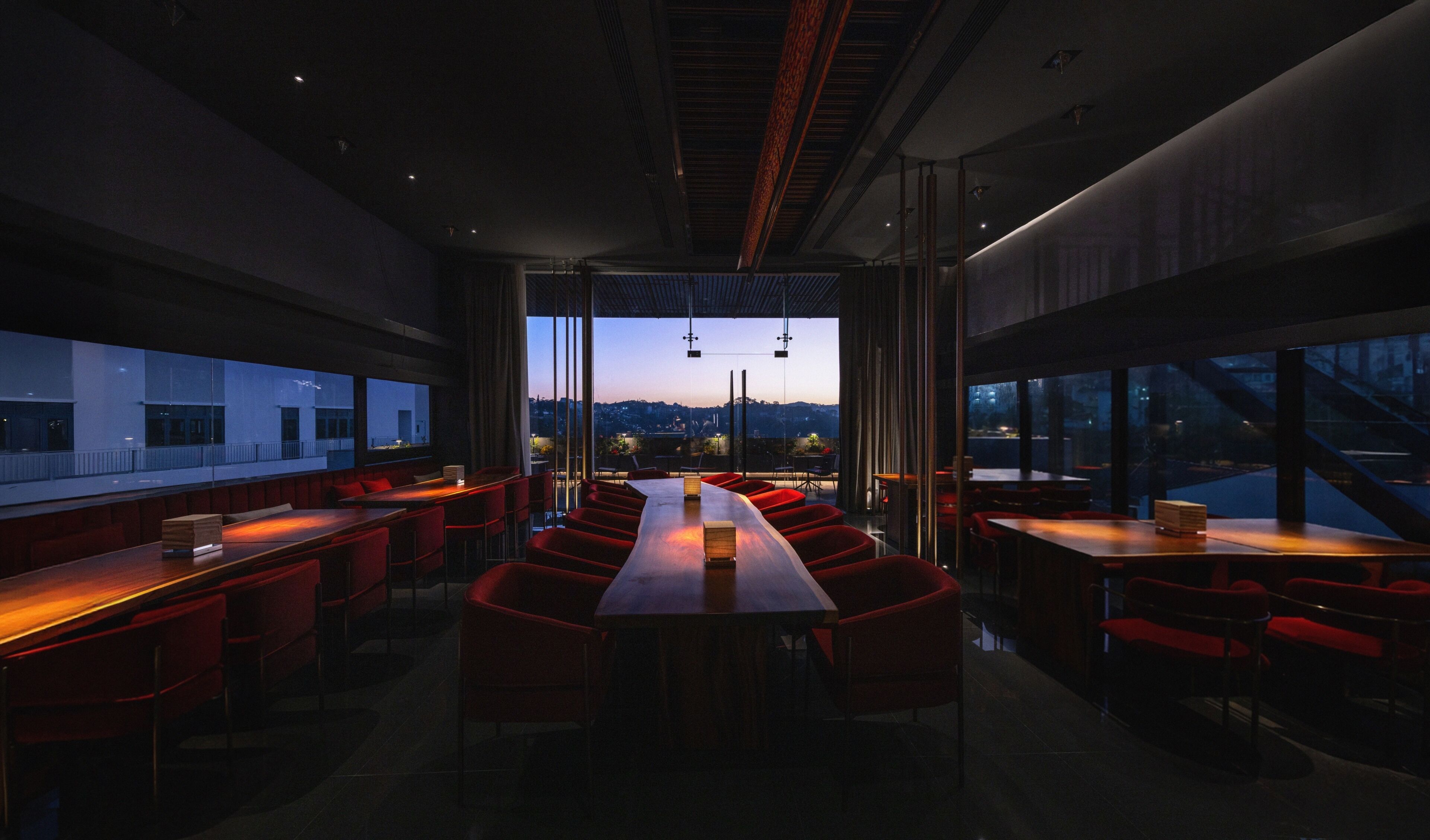 A modern dining area with a long table and red chairs, large windows, and a view of the cityscape.