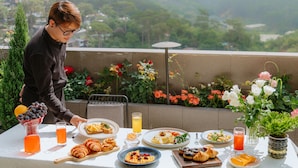 Une table dressée avec des aliments pour le petit-déjeuner, une personne qui sert la nourriture, et une vue sur la verdure et les bâtiments.