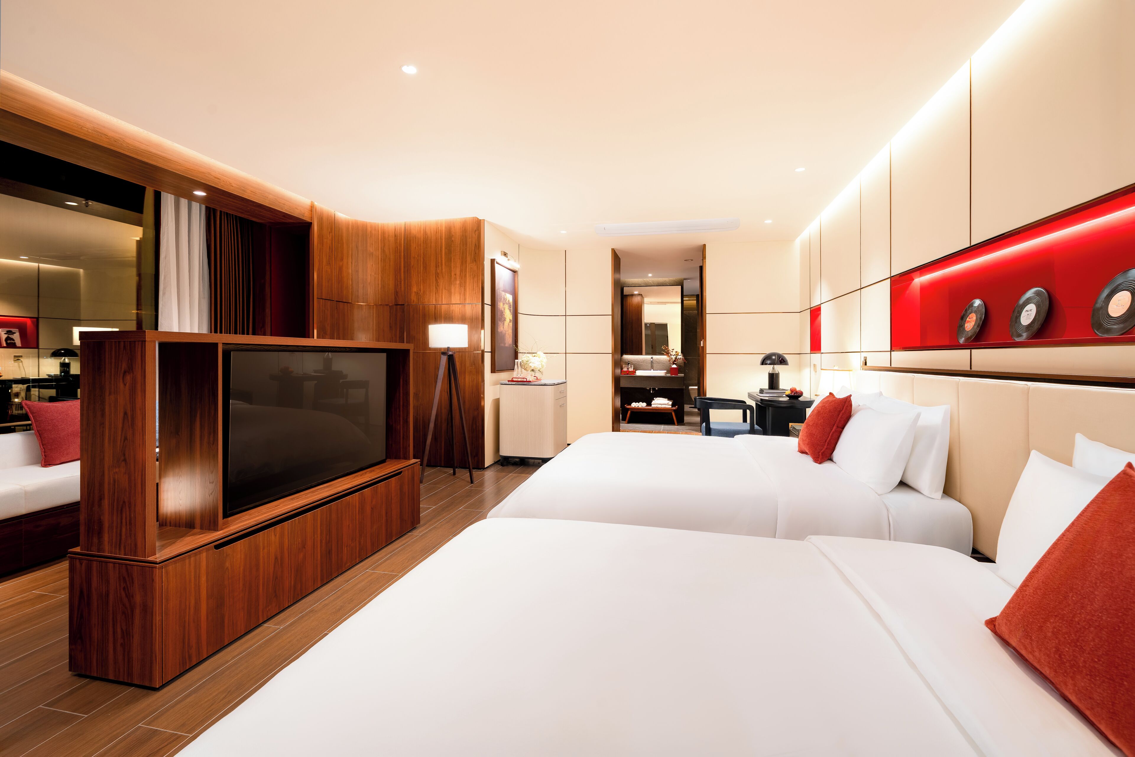 Une chambre d’hôtel moderne dotée d’un grand téléviseur à écran plat, de deux lits, d’un meuble de rangement en bois et d’un mur décoré de pochettes de disques rouges.
