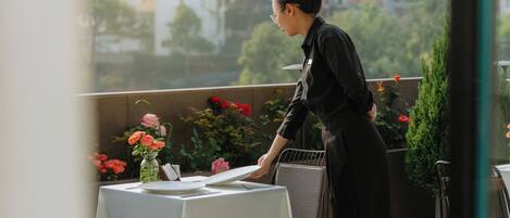 A woman setting up a table with a white tablecloth and a vase of flowers on a balcony overlooking a residential area.