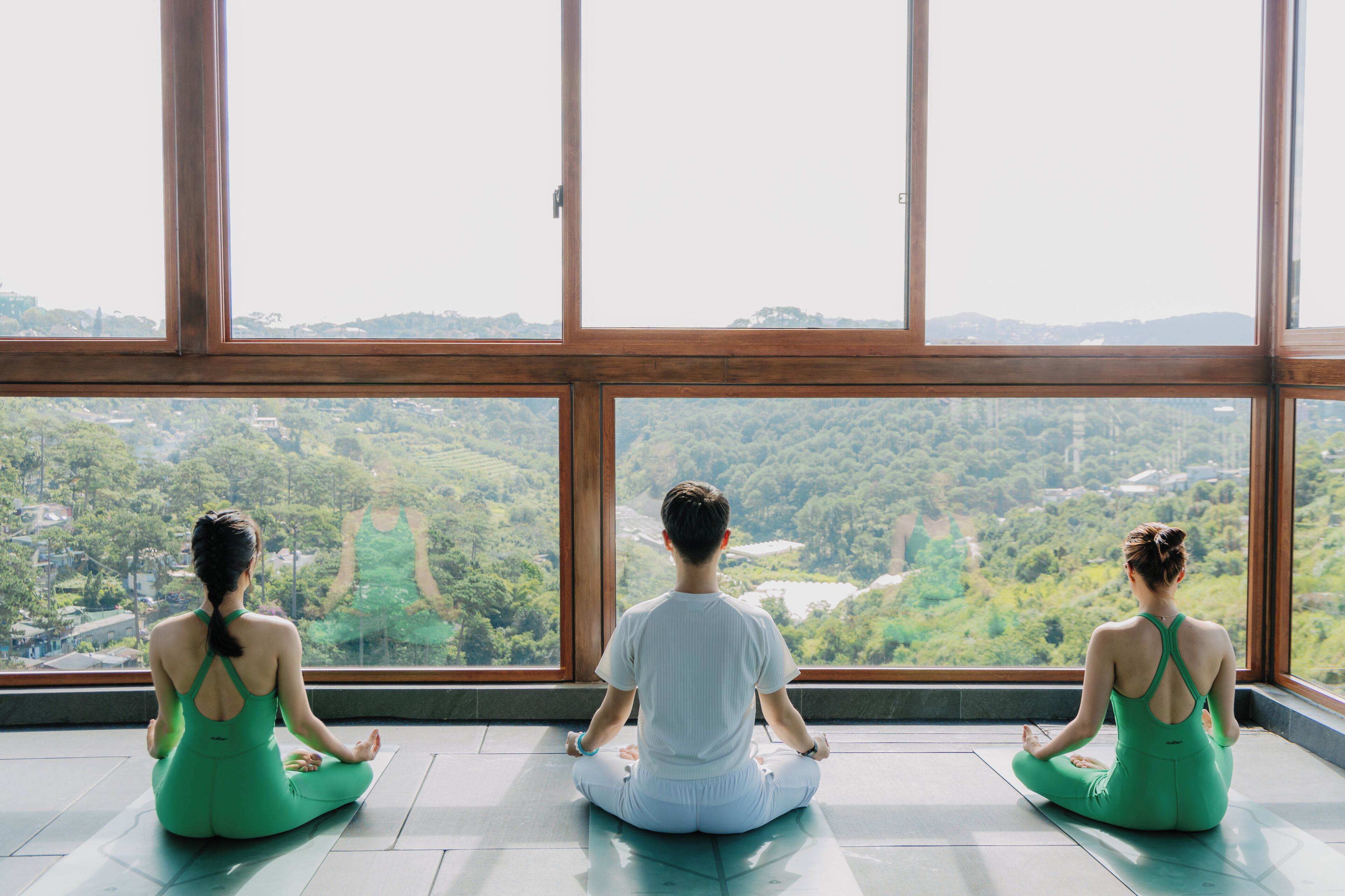 Three individuals seated in a meditative pose on a yoga mat inside a room with large windows.