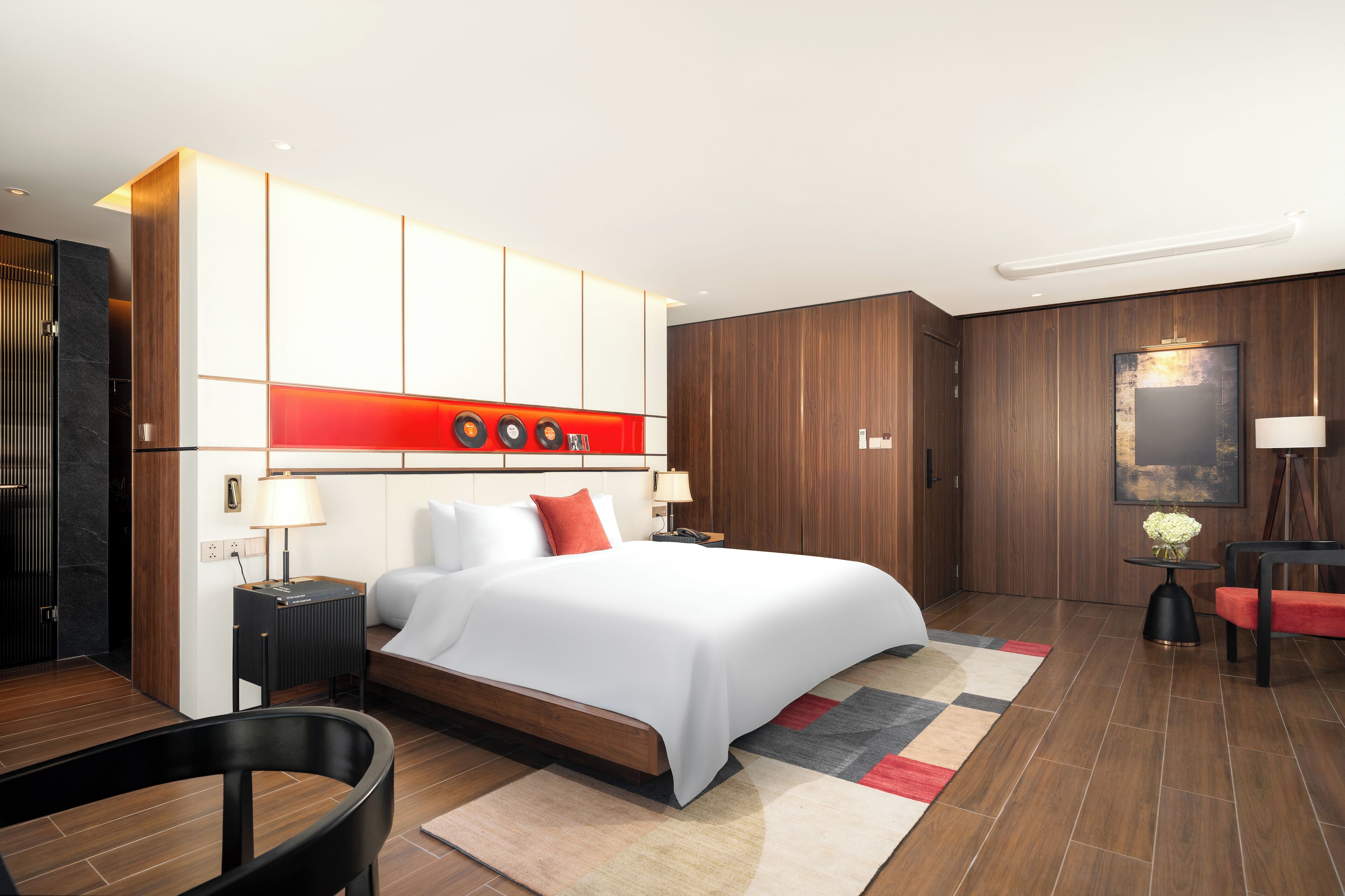 A modern hotel room with a large bed, wooden paneling, a red accent wall, and a small side table with a lamp.