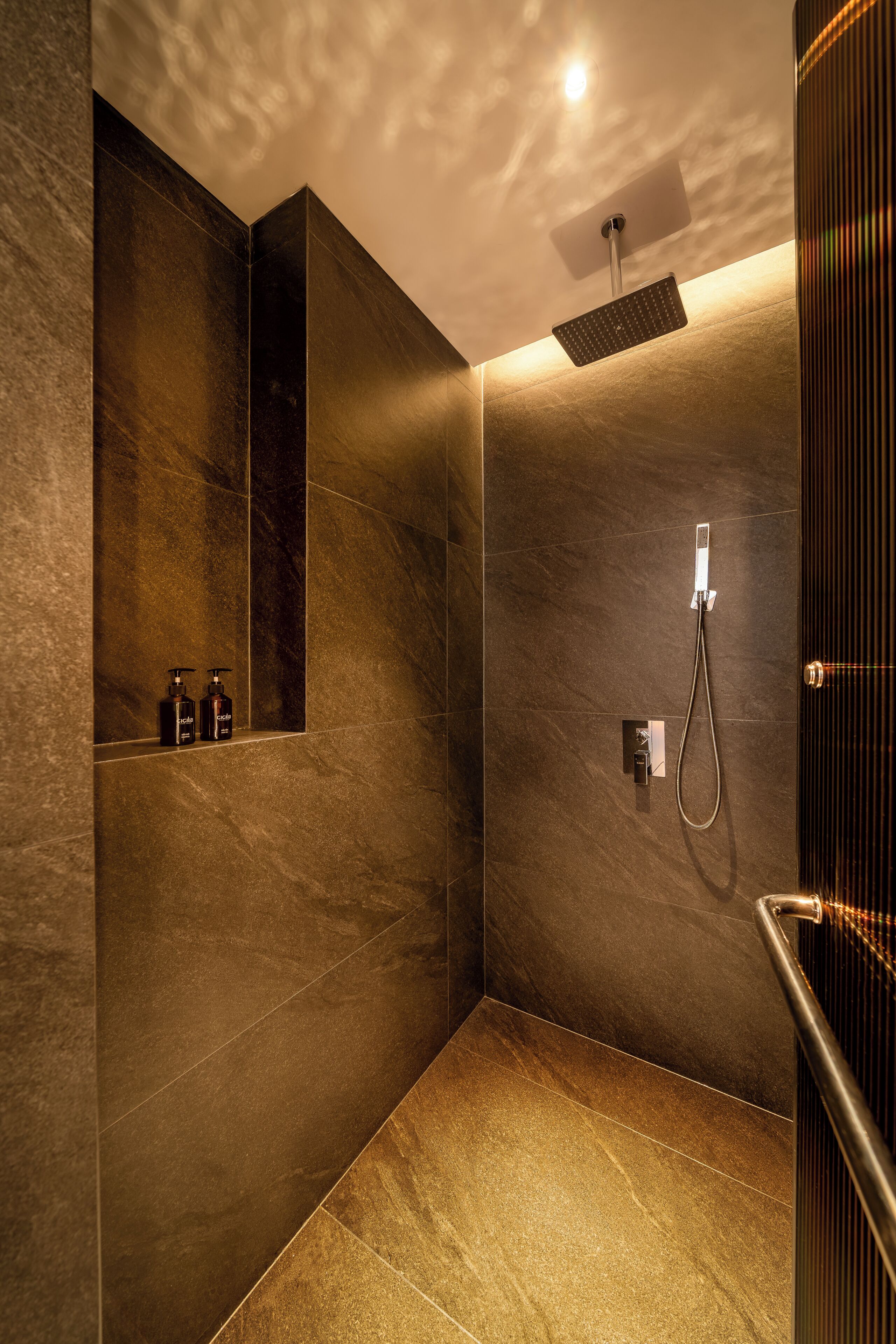 A modern shower area with a handheld showerhead, a control panel, and a small shelf with bottles.