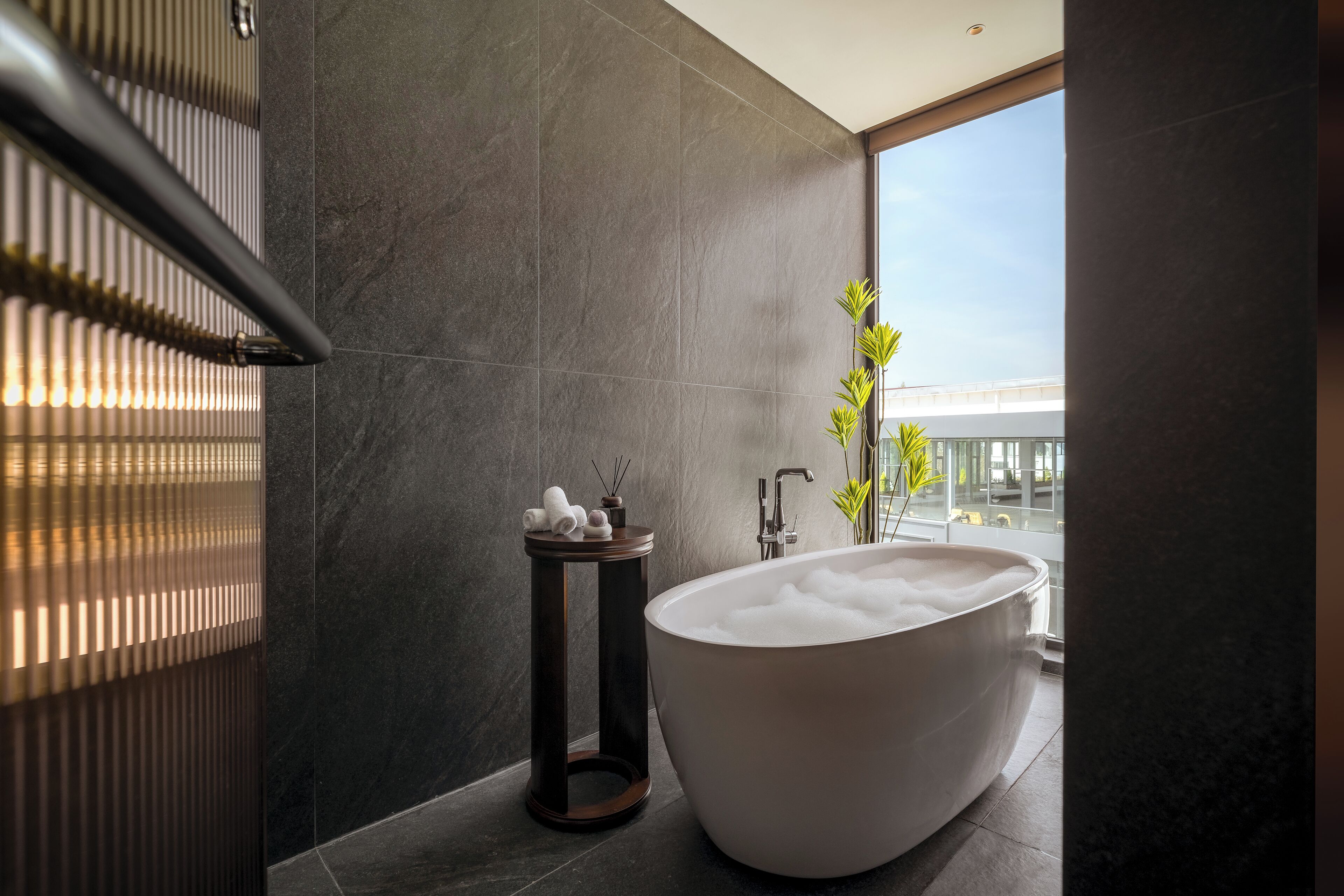 Een moderne badkamer met een vrijstaand bad, een klein nachtkastje met toiletartikelen, en uitzicht op een gebouw door een groot raam.