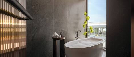 A modern bathroom with a freestanding bathtub, a small side table with toiletries, and a view of a building through a large window.