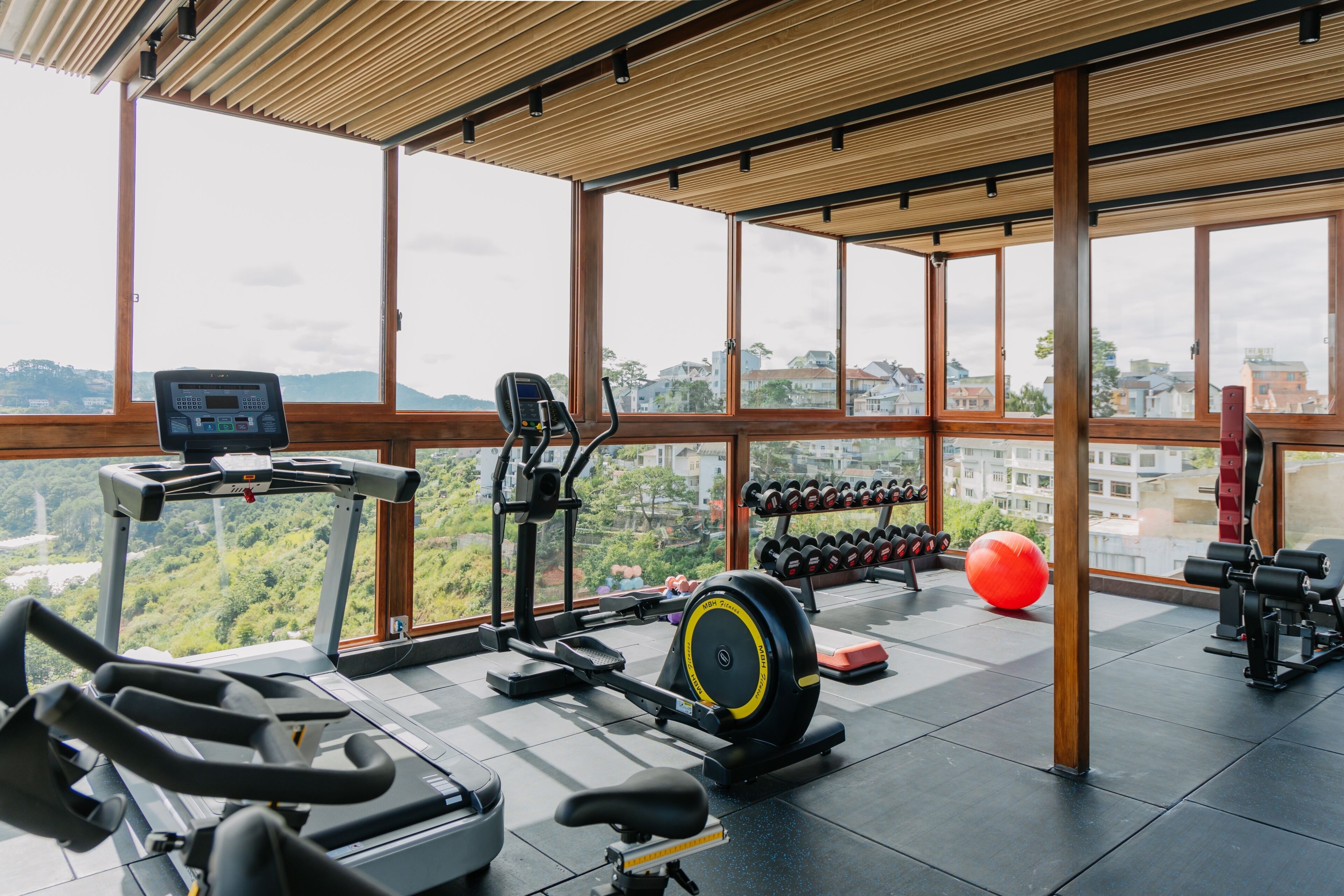 A gym with exercise equipment, large windows, and a view of greenery and buildings.