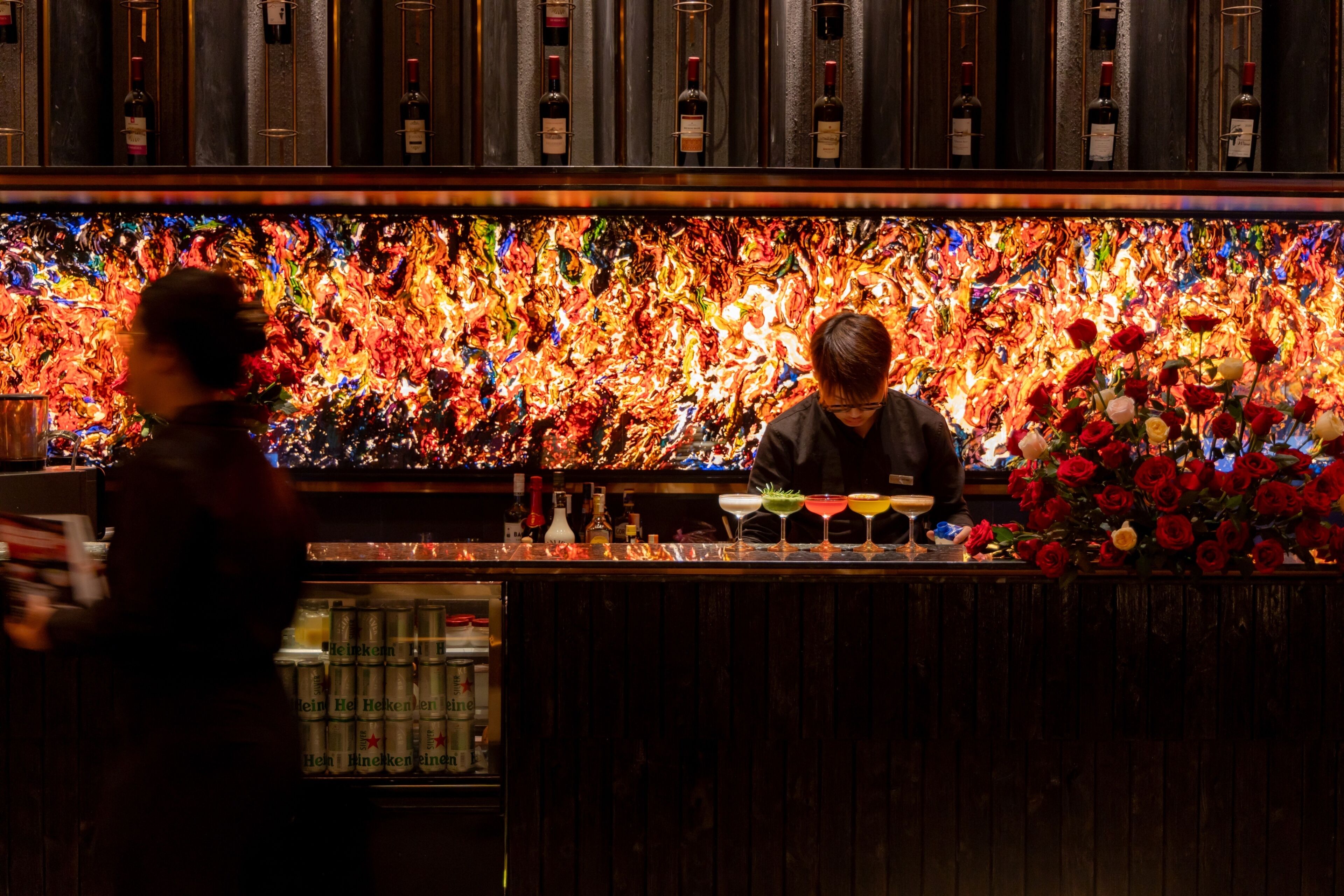 Eine Bar mit farbenfroher, abstrakter Wandgestaltung, alkoholischen Getränken und einem Barkeeper, der Cocktails zubereitet.