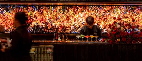 Eine Bar mit farbenfroher, abstrakter Wandgestaltung, alkoholischen Getränken und einem Barkeeper, der Cocktails zubereitet.