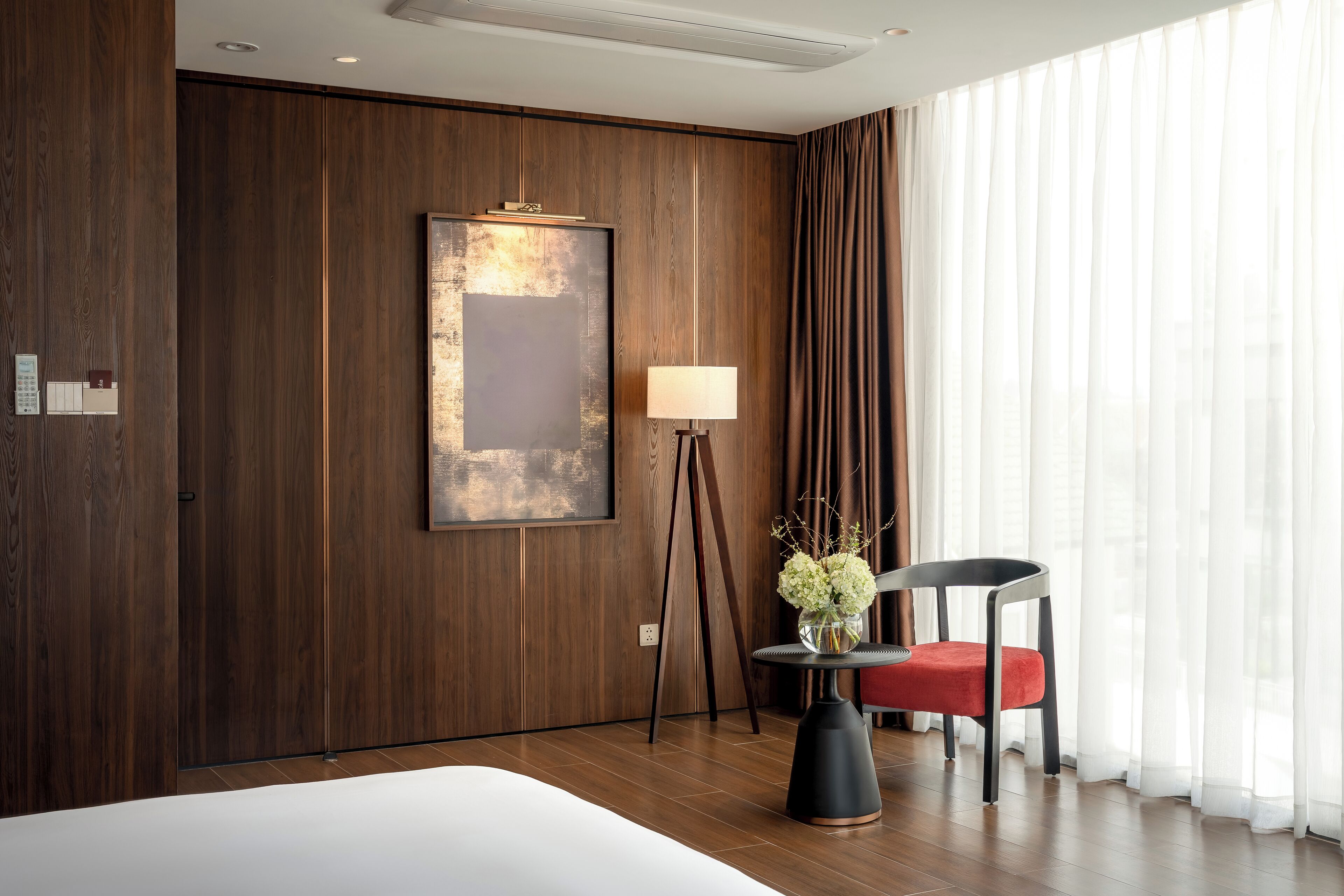Habitación de hotel con una cama grande, una lámpara de pie, una silla y vistas a través de cortinas traslúcidas.