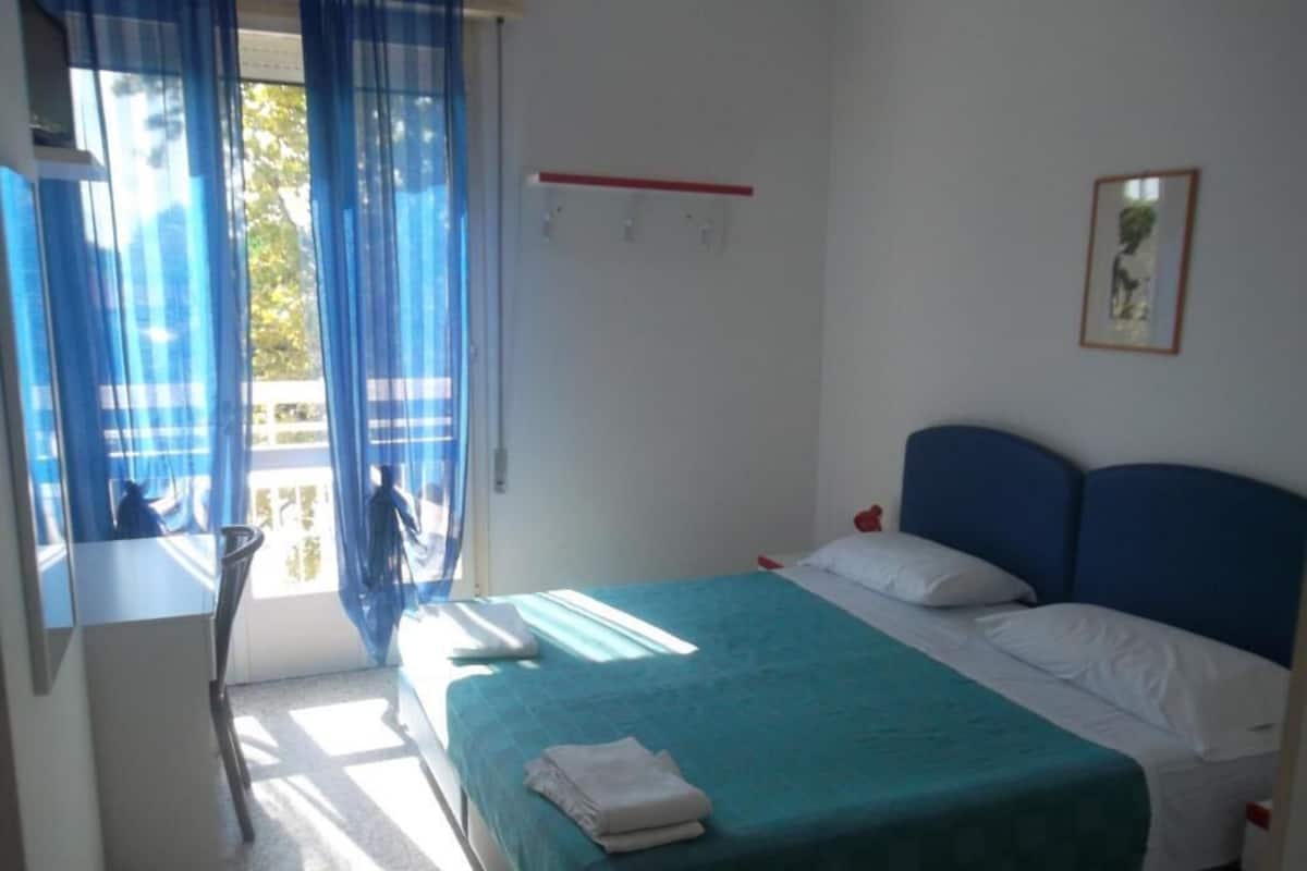 A bedroom with a bed, a window with curtains, a bedside table, and a wall-mounted shelf.