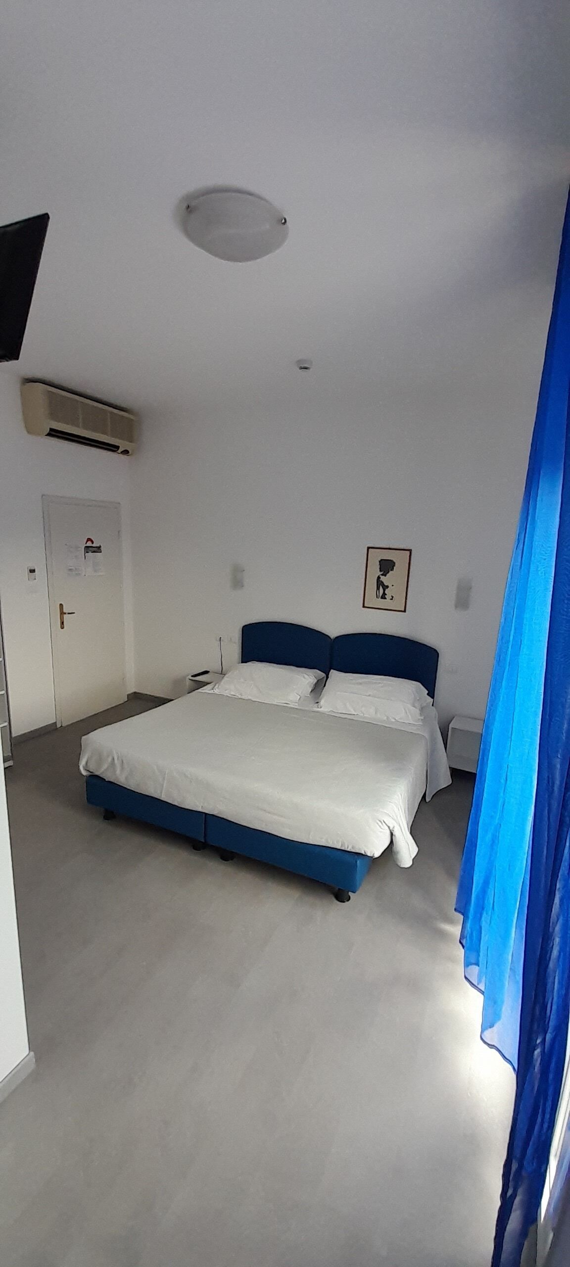 Une chambre à coucher comprenant un lit, une porte, un appareil de climatisation, un luminaire fixé au mur et un rideau.