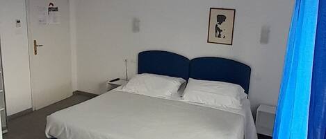 Une chambre à coucher comprenant un lit, une porte, un appareil de climatisation, un luminaire fixé au mur et un rideau.