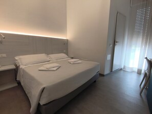A hotel room with a bed, bedside tables, a nightstand, a door with a window, and a chair. - Hotel Otello (Punta Marina)