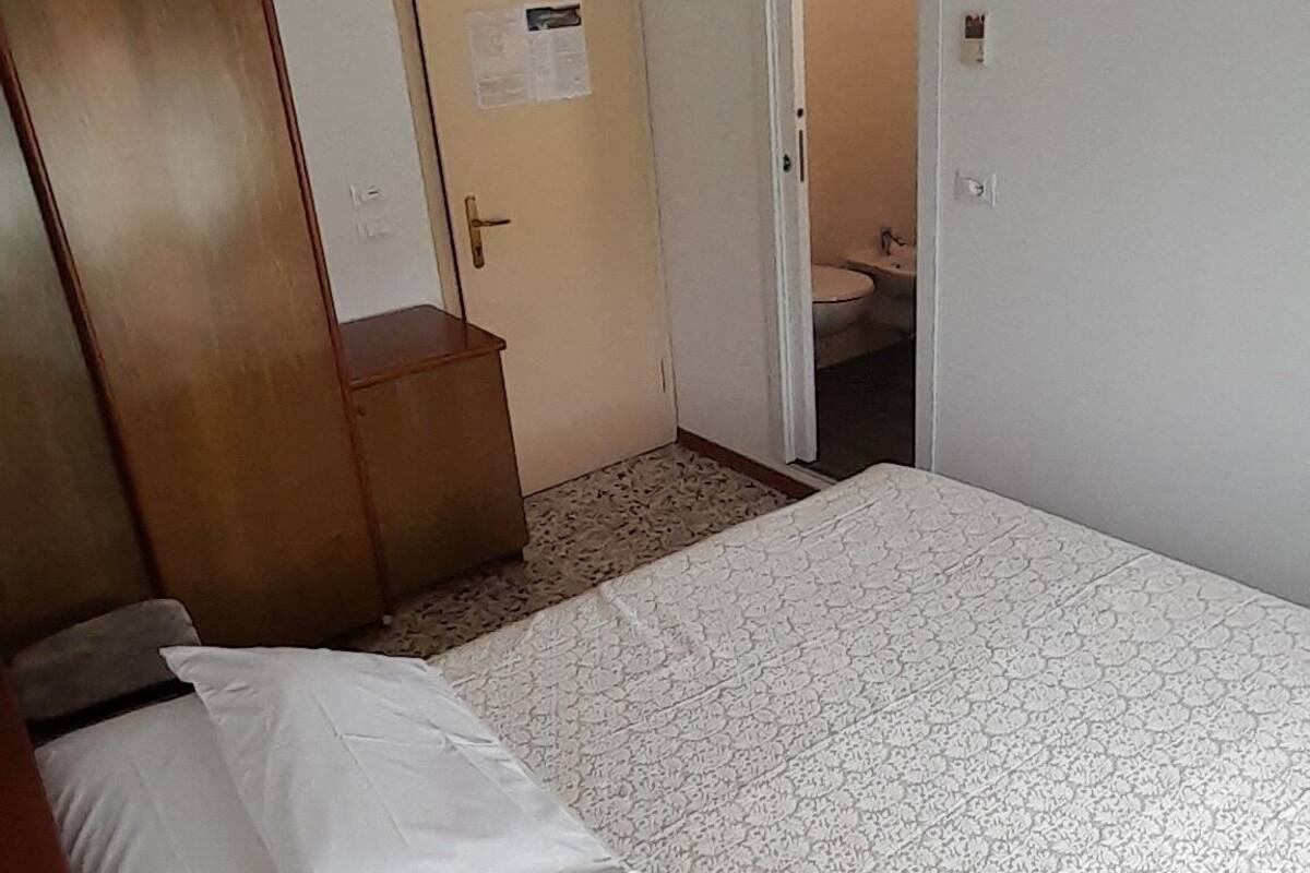 A bedroom with a bed, a ceiling fan, an air conditioning unit, a wooden wardrobe, and a door leading to a bathroom.