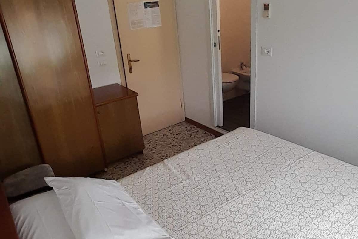A bedroom with a bed, a ceiling fan, an air conditioning unit, a wooden wardrobe, and a door leading to a bathroom.