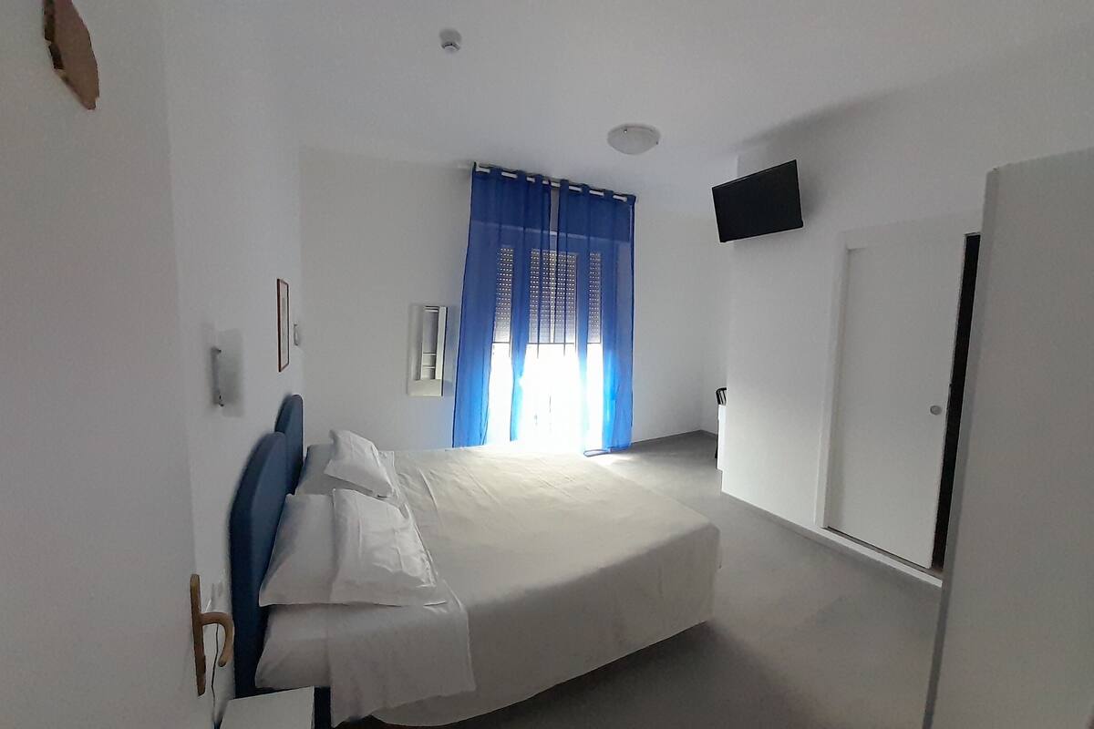 A bedroom with a bed, a door, a wall-mounted TV, and a window with curtains.