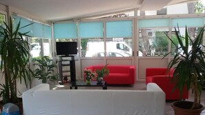 A room with a white sofa, red chairs, a TV on a stand, and potted plants. - Hotel Otello (Punta Marina)