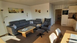 Ein Wohnzimmer mit einer Couch, einem Couchtisch und einem Essbereich mit Stühlen. Im Hintergrund ist eine Küche zu sehen.