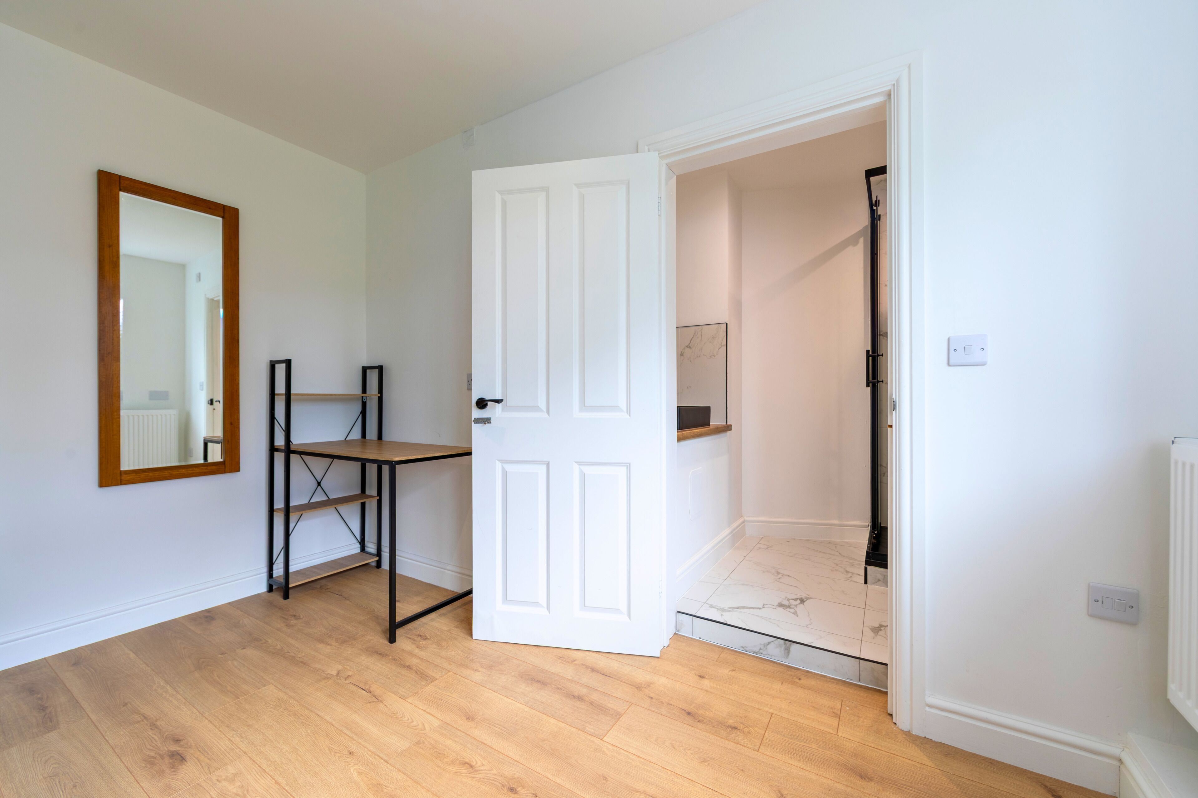 A room with a white door, a wooden floor, a metal shelf, and a mirror on the wall.