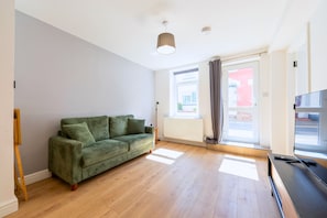 Une pièce avec un canapé vert, un plancher en bois, un radiateur, une fenêtre avec des rideaux et un téléviseur posé sur un meuble.