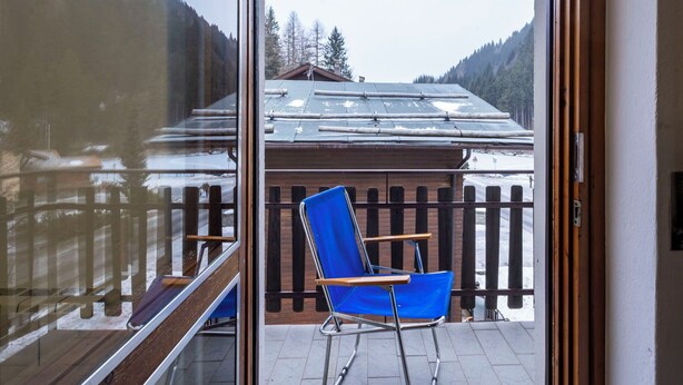 A balcony with a blue chair, wooden railing, and a view of a mountain and a building with a sloped roof. - Cosy apartment for 4 people with WIFI, TV, balcony and pets allowed (Scalcoggia)