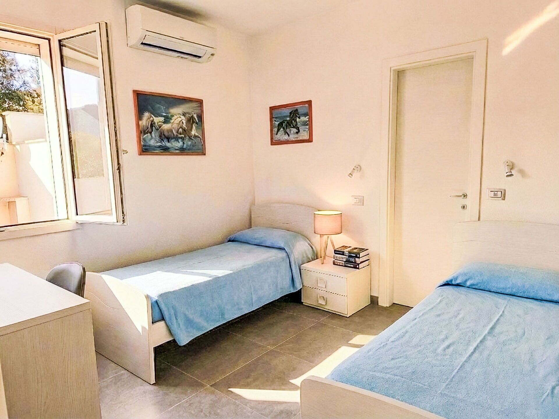 Een slaapkamer met twee eenpersoonsbedden, een bureau, een stoel, een airconditioner en twee ingelijste foto's aan de muur.
