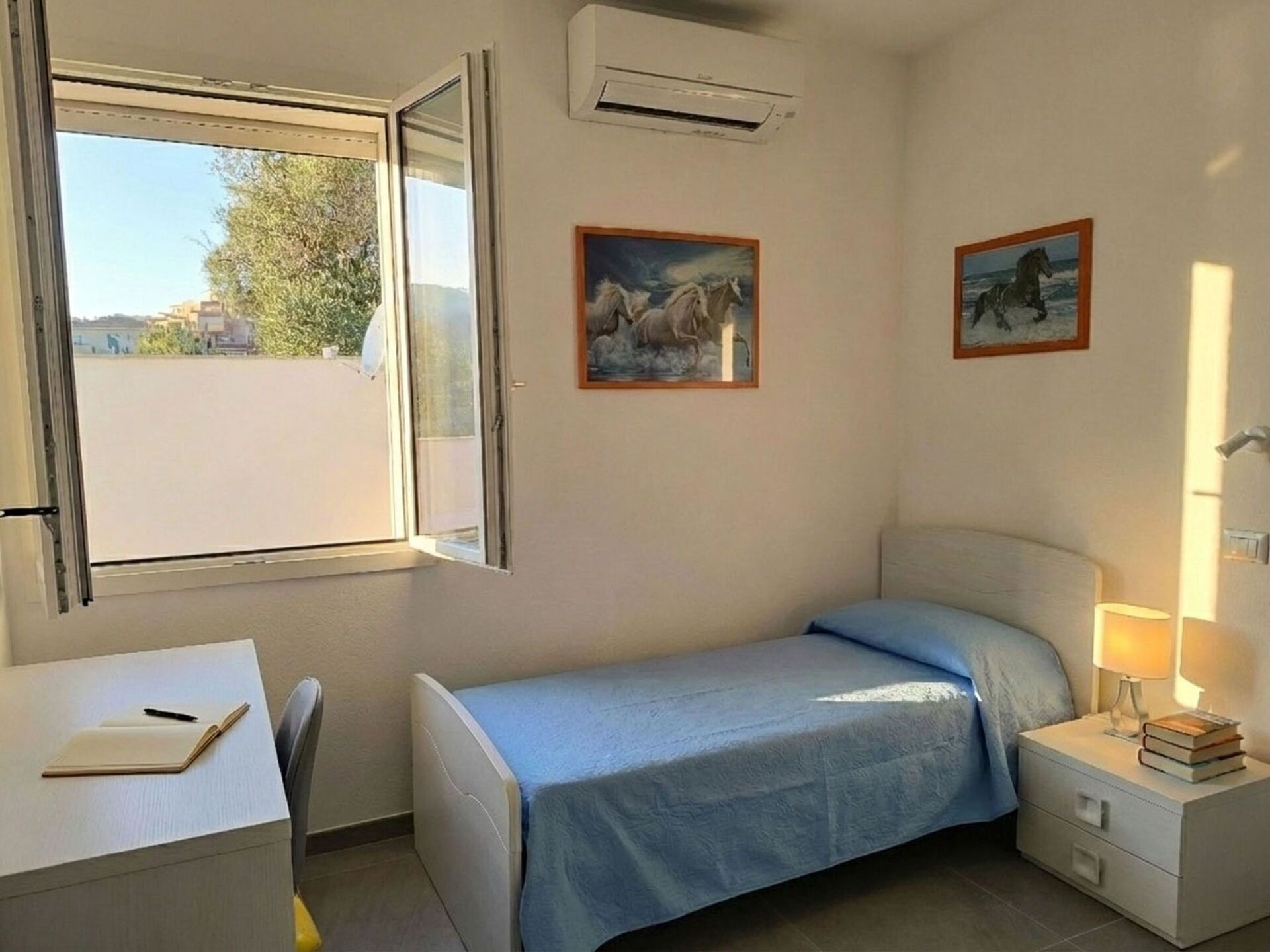 Une petite pièce propre, avec un lit, un bureau et une fenêtre. Il y a deux tableaux accrochés au mur et un climatiseur au-dessus du lit.