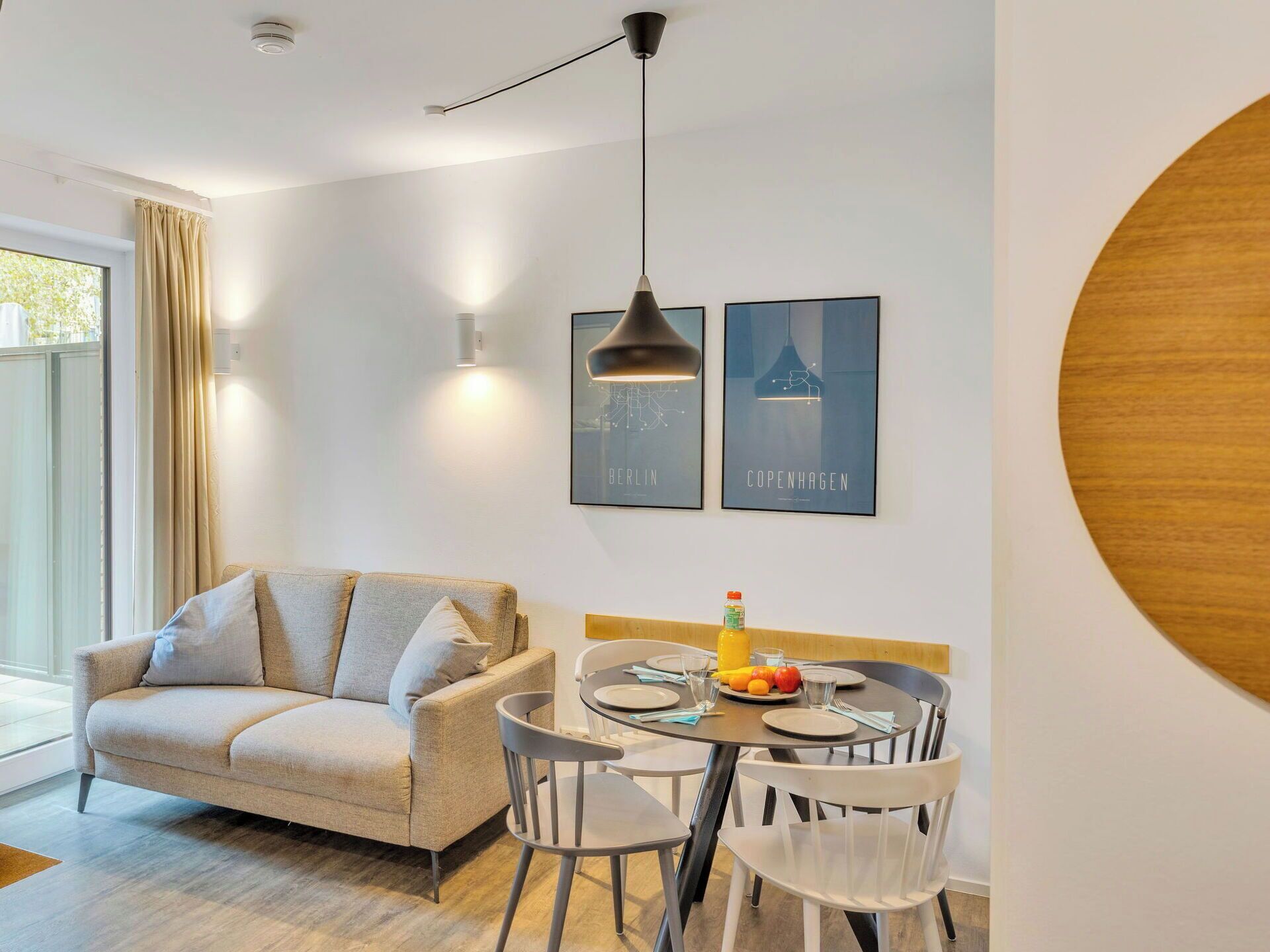 Una sala de estar moderna con un sofá, una mesa de comedor y sillas. Hay dos cuadros en la pared, una lámpara colgante y un elemento decorativo de madera grande.