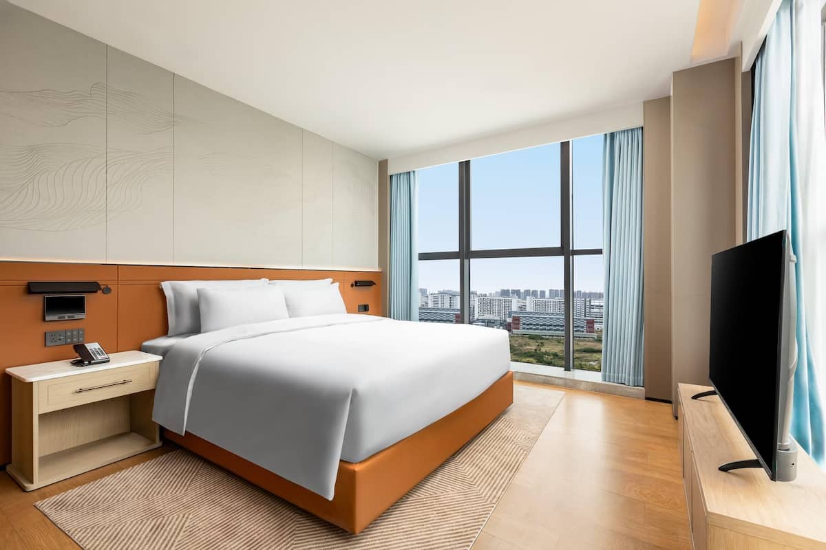 Habitació d'hotel moderna amb un llit gran, una televisió sobre un suport de fusta, una nit falda amb un telèfon i vista a la ciutat a través de grans finestres.