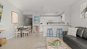 A modern living room with a grey sofa, a coffee table with a geometric pattern, and a dining area with a wooden table and chairs. - Gulfside 107 at Destin West ~ Ground Floor  ~ Direct Pool & Beach Access (Fort Walton Beach)
