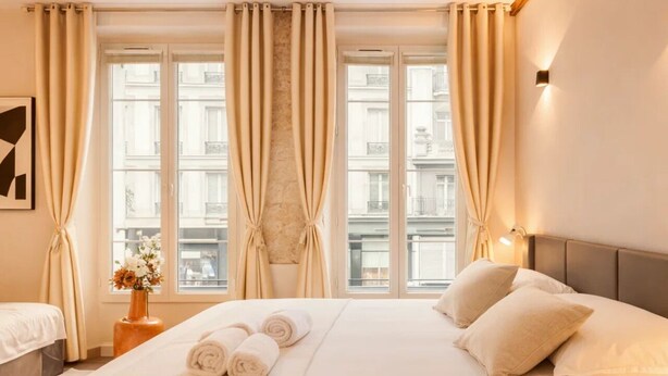 A hotel room with a large bed, two bedside tables, a vase with flowers, and a view of buildings through the windows. - 1528 Nazareth (Paris)