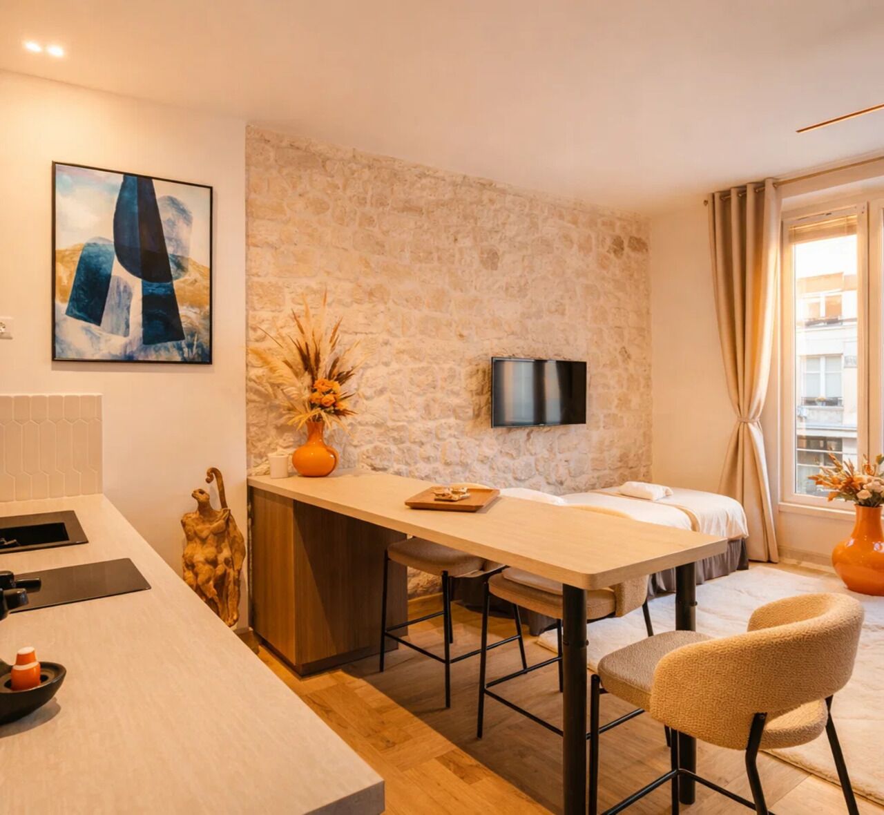 A modern hotel room with a stone wall, a bed, a desk with chairs, a TV, and a window with curtains.