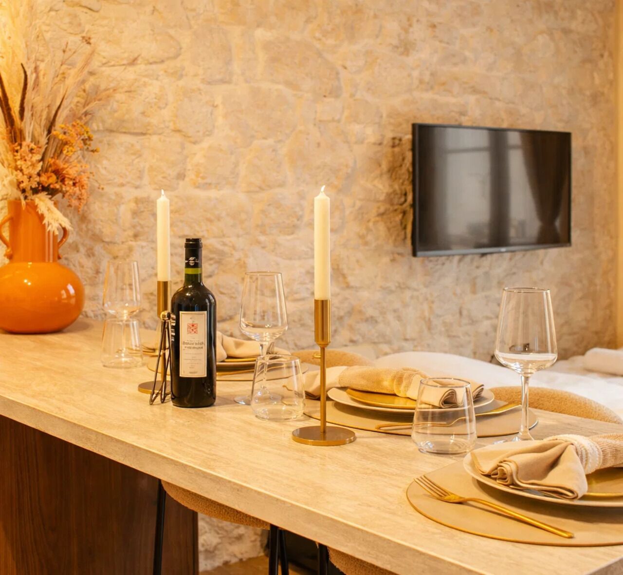 A dining table set with candles, wine glasses, and plates, with a stone wall and a flat-screen TV in the background.
