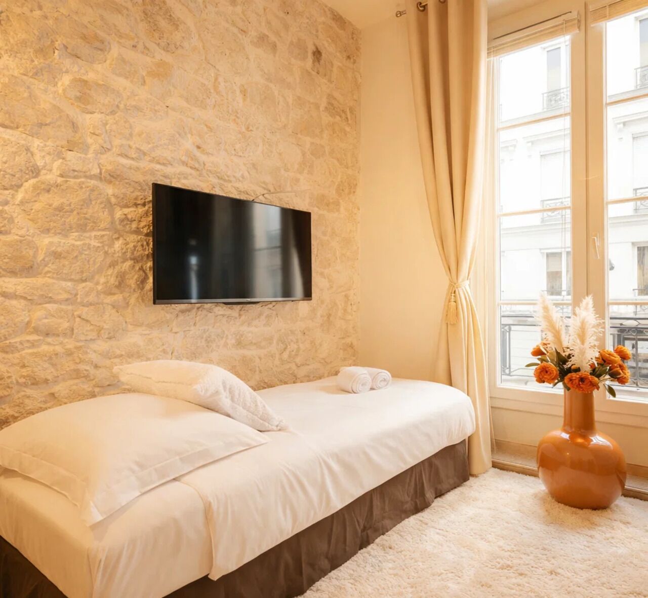 Un dormitorio con pared de piedra, un televisor de pantalla plana sobre la cama, una ventana con cortinas y un florero con flores en el alféizar.