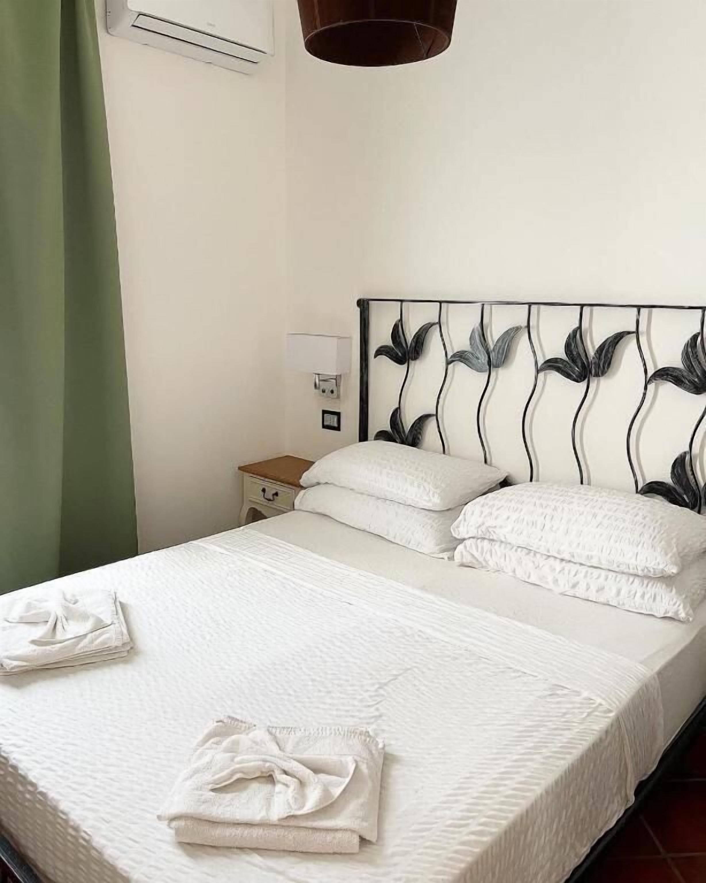 Una cama bien hecha con sábanas y almohadillas blancas, una mesita de noche con lámpara y un aire acondicionado de pared.